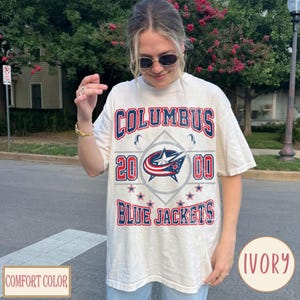 Könnte beinhalten: Ein elfenbeinfarbenes Kurzarm-T-Shirt mit einem verwaschenen Grafikdruck, der den Text COLUMBUS BLUE JACKETS, das Jahr 2000 und ein zentrales Stern- und Bandlogo in Rot, Weiß und Blau zeigt. Das Design enthält kleine Sternakzente.