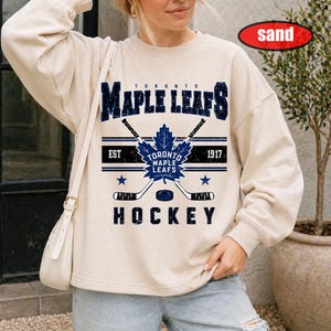 May include: A sand-colored crewneck sweatshirt featuring a distressed blue graphic of the Toronto Maple Leafs logo, two crossed hockey sticks, and the text Toronto Maple Leafs Est 1917 Hockey. The design includes two small blue stars.