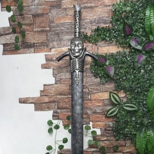 May include: A large, decorative sword with a skull-shaped hilt and a skeletal ribcage design. The blade is a dark gray color, and the sword is displayed against a brick wall with green foliage.
