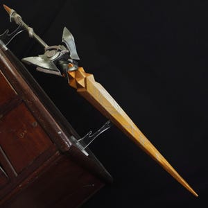 May include: A decorative sword with a wooden handle and a silver and gold blade. The handle is a light brown color with a geometric design. The blade has a silver finish with gold accents. The sword is displayed on a clear stand against a dark background.