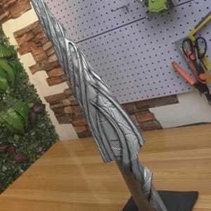 May include: A silver-colored, stylized sculpture with a textured, leaf-like design. The sculpture is mounted on a wooden base and a black square stand. The artwork has a sharp, pointed tip and a curved, flowing form.