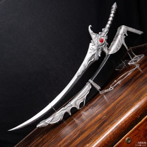May include: A silver fantasy sword with intricate dragon-inspired details and a red gem set in the hilt. The curved blade is displayed with its scabbard and a decorative guard. The sword is made of metal and is on a wooden surface.