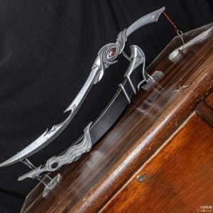 May include: Two fantasy-style silver daggers with ornate designs and a red gem. One dagger is sheathed in a black scabbard. The daggers are displayed on a wooden surface against a black backdrop.