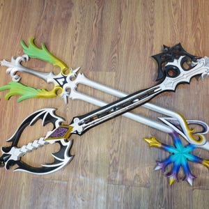 May include: Three decorative keyblades with intricate designs. One keyblade features a black and white handle with a purple accent, another has green and gold accents, and the third has blue and gold accents. A small charm is attached to one.