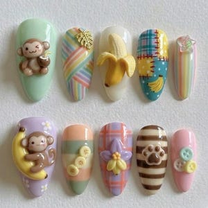May include: A set of ten almond-shaped artificial nails with various designs. The designs include a monkey with a coconut, a banana, plaid patterns, stripes, and a paw print. Colors include pastel shades of green, purple, pink, yellow, and brown.