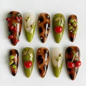 May include: A set of ten almond-shaped, press-on nails with various designs. Some nails feature a brown tortoiseshell pattern, while others have green tips and are adorned with cherries, olives, and cocktail glasses. Some nails have gold accents.