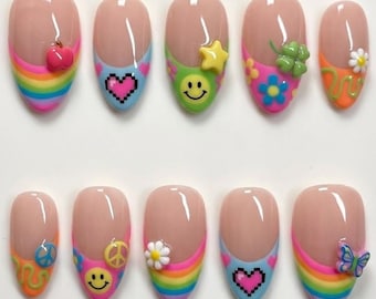 Könnte beinhalten: Ein Set aus zehn künstlichen Nägeln mit hellrosa Grundierung und farbenfrohen Designs. Jeder Nagel hat eine regenbogenfarbene French-Spitze und verschiedene 3D-Verzierungen, darunter ein Herz, ein Smiley, ein Stern und ein Schmetterling. Die Nägel sind in zwei Reihen angeordnet.