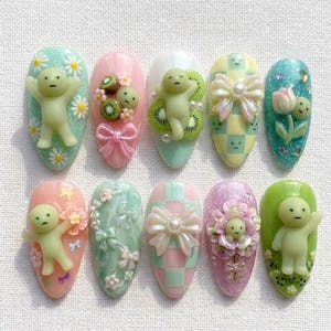 May include: A collection of ten almond-shaped nail designs. Each nail features a unique design, including 3D figures, bows, flowers, and kiwi fruit. The color palette includes pastel shades of pink, green, blue, and yellow, with glitter accents.