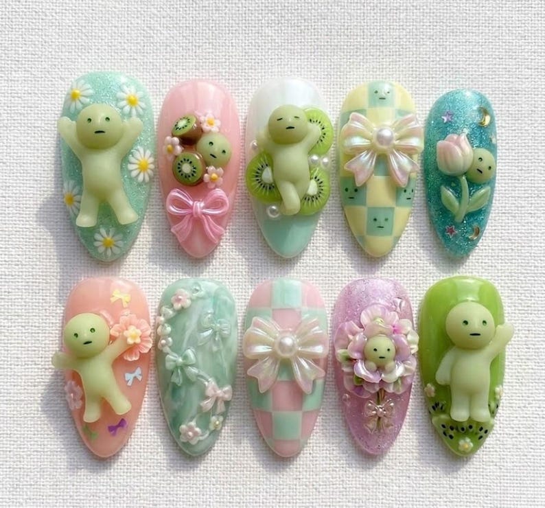 May include: A collection of ten almond-shaped nail designs. Each nail features a unique design, including 3D figures, bows, flowers, and kiwi fruit. The color palette includes pastel shades of pink, green, blue, and yellow, with glitter accents.