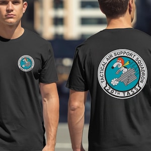 May include: Black t-shirts with a circular graphic on the front and back. The graphic features a cartoon dog in a red helmet and scarf, holding a gun, with the text "TACTICAL AIR SUPPORT SQUADRON" and "20TH TASS" in a circle.