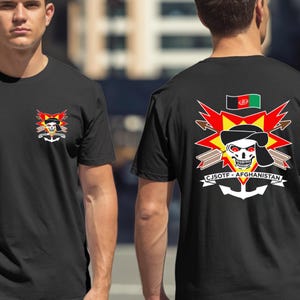 Può includere: T-shirt nere con un design grafico raffigurante un teschio con bandana, un'esplosione rossa e gialla e la scritta "CJSOTF-AFGHANISTAN". Il design include un'ancora e la bandiera dell'Afghanistan.