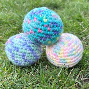 May include: Three colorful crocheted balls rest on a bed of green grass. The balls are in shades of blue, green, pink, and yellow. The top ball is teal with pink and purple accents. The middle ball is blue and green. The bottom ball is pastel.