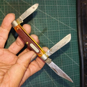 May include: A multi-blade pocket knife featuring three silver-toned steel blades in various positions. The handle is constructed with amber-colored translucent scales over a dark brown core, accented by brass pins and a central metal shield inlay.