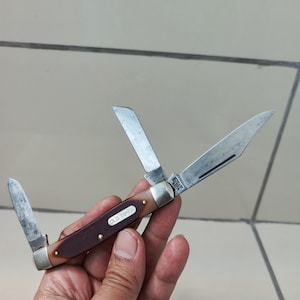 May include: A folding pocket knife with three silver-toned metal blades and a brown handle featuring a white inlay with the text OLD TIMER. The handle is secured with small brass rivets and has metallic bolsters at both ends of the knife frame.