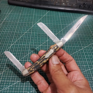 May include: A folding pocket knife with three silver-toned metal blades extended at different angles. The handle features a textured, mottled brown and tan pattern with small brass rivets and a central metal inlay shield.