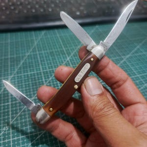Peut inclure: Un couteau de poche pliant avec trois lames en acier argenté déployées. Le manche est marron avec une surface texturée et comporte une incrustation en métal argenté avec le texte OLD TIMER. La base de la lame porte l'inscription ULSTER USA.