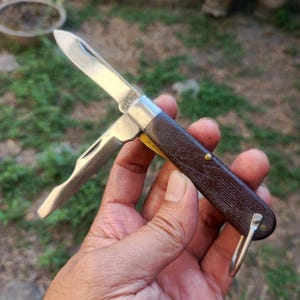 Può includere: Un coltellino tascabile pieghevole con manico testurizzato marrone scuro e due lame in metallo color argento estratte. Il coltello presenta un piccolo anello in metallo all'estremità del manico e rivetti metallici visibili sul corpo.