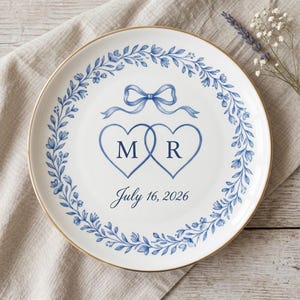May include: White ceramic plate with a gold rim, featuring a blue floral wreath design. The plate has two intertwined heart shapes with the letters "M" and "R", a blue bow, and the date "July 16, 2026".