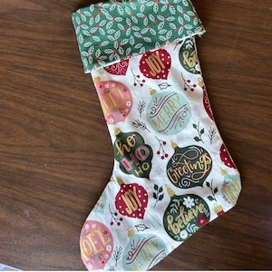 May include: A white Christmas stocking with a green and white cuff. The stocking is decorated with red, green, and gold ornaments with festive sayings like "Joy", "Merry", "Greetings", and "Believe".