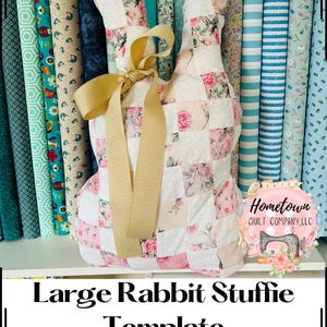 May include: A large pink and white patchwork rabbit stuffed animal with a brown bow. The rabbit is sitting on a shelf with a variety of fabric rolls behind it. The text "Hometown Quilt Company, LLC" is visible in the image.