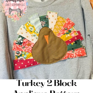 Könnte beinhalten: Ein grauer Pullover mit einer Patchwork-Truthahn-Applikation. Der Truthahn ist aus verschiedenen Stoffen in Rot-, Rosa-, Gelb-, Grün- und Brauntönen gefertigt. Der Text "Turkey 2 Block Applique Pattern Pieces" ist unter dem Bild gedruckt.