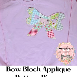 May include: A pink sweatshirt featuring a patchwork fabric bow applique. The bow is made from various floral and checked patterns in shades of pink, blue and green. The sweatshirt has the text "Hometown Quilt Company, LLC" printed in the bottom right corner.