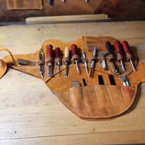 Pode incluir: Um rolo de ferramentas de couro castanho claro, aberto e plano, exibindo v&aacute;rias ferramentas de trabalho em couro. O rolo &eacute; de cor castanho claro, com uma correia para prend&ecirc;-lo. As ferramentas incluem cinz&eacute;is, pun&ccedil;&otilde;es e outros implementos com cabos de madeira.