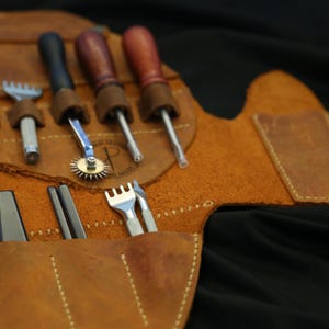 Pode incluir: Um kit de ferramentas de artesanato em couro com v&aacute;rias ferramentas, incluindo furadores, um sulcador de costura, um cortador rotativo e uma ferramenta em forma de garfo. As ferramentas s&atilde;o guardadas em um estojo enrol&aacute;vel de couro marrom com um design costurado. O fundo &eacute; preto.