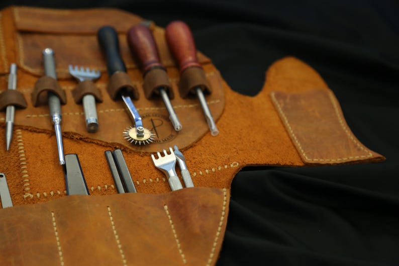 Pode incluir: Um kit de ferramentas de artesanato em couro com v&aacute;rias ferramentas, incluindo furadores, um sulcador de costura, um cortador rotativo e uma ferramenta em forma de garfo. As ferramentas s&atilde;o guardadas em um estojo enrol&aacute;vel de couro marrom com um design costurado. O fundo &eacute; preto.