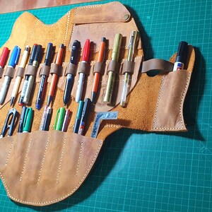 Pode incluir: Um estojo de canetas de couro castanho claro feito &agrave; m&atilde;o com v&aacute;rias canetas e marcadores de v&aacute;rias cores. O estojo est&aacute; aberto, mostrando os instrumentos de escrita em ranhuras e bolsos individuais. Uma correia de couro prende o estojo quando fechado. O fundo &eacute; uma base de corte verde.