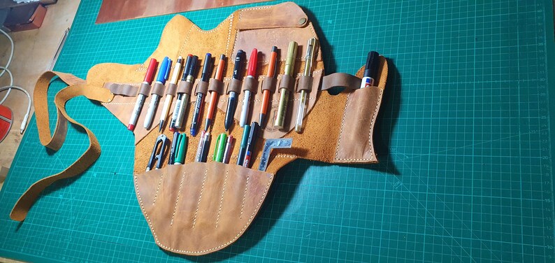 Pode incluir: Um estojo de canetas de couro castanho claro feito &agrave; m&atilde;o com v&aacute;rias canetas e marcadores de v&aacute;rias cores. O estojo est&aacute; aberto, mostrando os instrumentos de escrita em ranhuras e bolsos individuais. Uma correia de couro prende o estojo quando fechado. O fundo &eacute; uma base de corte verde.