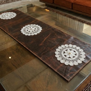 May include: A brown leather table runner featuring three white lace doilies. The runner is rectangular and placed on a glass-topped coffee table. The doilies have a floral design.