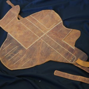 Pode incluir: Uma almofada de sela de couro castanho com costuras brancas e uma longa correia. A almofada tem uma forma &uacute;nica e foi concebida para proteger as costas do cavalo. O couro tem um aspeto envelhecido.