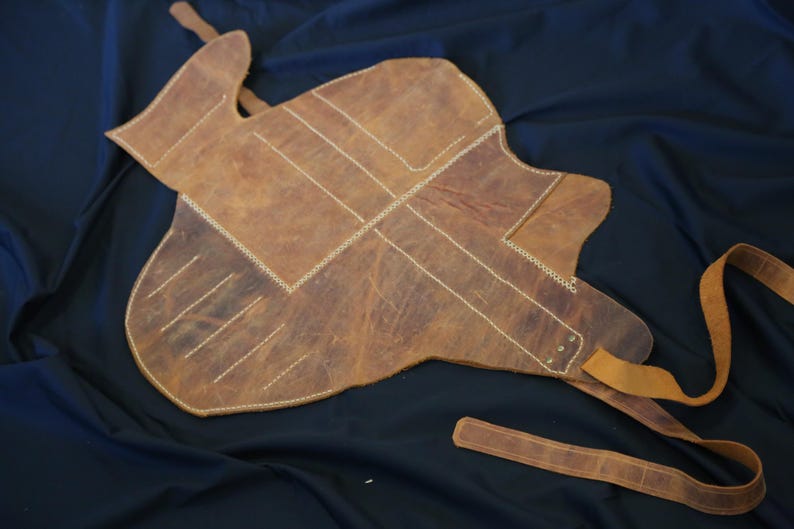 Pode incluir: Uma almofada de sela de couro castanho com costuras brancas e uma longa correia. A almofada tem uma forma &uacute;nica e foi concebida para proteger as costas do cavalo. O couro tem um aspeto envelhecido.