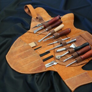 Pode incluir: Um kit de ferramentas de artesanato em couro com v&aacute;rias ferramentas, incluindo cinz&eacute;is, pun&ccedil;&otilde;es e um sulcador de costura. As ferramentas t&ecirc;m cabos de madeira e s&atilde;o fixadas em uma caixa de couro castanho claro com uma al&ccedil;a.