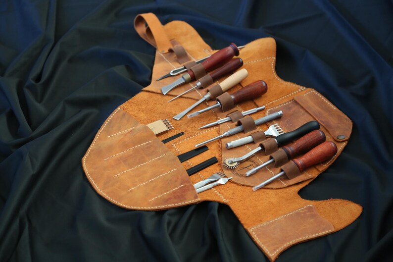 Pode incluir: Um kit de ferramentas de artesanato em couro com v&aacute;rias ferramentas, incluindo cinz&eacute;is, pun&ccedil;&otilde;es e um sulcador de costura. As ferramentas t&ecirc;m cabos de madeira e s&atilde;o fixadas em uma caixa de couro castanho claro com uma al&ccedil;a.