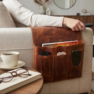 May include: A brown leather couch organizer with multiple pockets, holding books, a phone, and other items. The organizer is attached to the arm of a beige couch. A cup of coffee, glasses, and a book are on a nearby table.