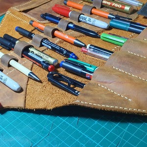 Pode incluir: Um estojo de caneta de couro marrom aberto, com canetas e marcadores de v&aacute;rias cores. O estojo tem presilhas individuais para prender as canetas. O couro tem uma apar&ecirc;ncia texturizada com costuras brancas.