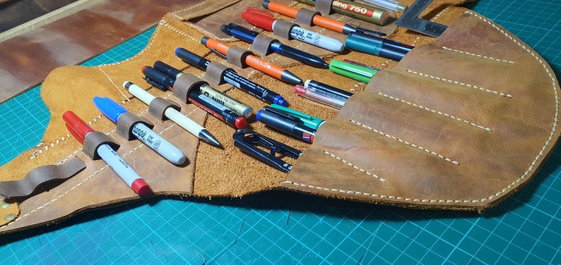 Pode incluir: Um estojo de caneta de couro marrom aberto, com canetas e marcadores de v&aacute;rias cores. O estojo tem presilhas individuais para prender as canetas. O couro tem uma apar&ecirc;ncia texturizada com costuras brancas.