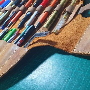 Pode incluir: Um estojo de canetas de couro castanho claro, aberto e preenchido com v&aacute;rias canetas e marcadores. O estojo possui v&aacute;rios compartimentos e uma al&ccedil;a. As costuras s&atilde;o vis&iacute;veis. O estojo est&aacute; sobre uma superf&iacute;cie verde.