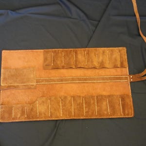 May include: A brown leather tool roll with multiple pockets and a strap for securing. The roll is rectangular when open, with white stitching and a small pocket on the left side. The leather has a textured appearance.