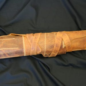 Pode incluir: Uma bainha de faca de couro marrom enrolada, com fecho de aba. A bainha &eacute; presa com v&aacute;rias tiras de couro enroladas ao redor do corpo. Costuras brancas acentuam as bordas e costuras, criando uma apar&ecirc;ncia r&uacute;stica e artesanal.