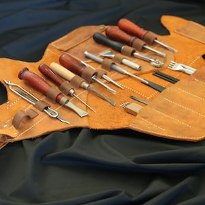 Pode incluir: Um kit de ferramentas de artesanato em couro com v&aacute;rias ferramentas, incluindo cinz&eacute;is, furadores e vincadores de bordas. As ferramentas t&ecirc;m cabos de madeira e s&atilde;o armazenadas em um estojo enrol&aacute;vel de couro castanho. O estojo est&aacute; aberto, mostrando as ferramentas em um fundo preto.