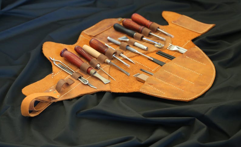 Pode incluir: Um kit de ferramentas de artesanato em couro com v&aacute;rias ferramentas, incluindo cinz&eacute;is, furadores e vincadores de bordas. As ferramentas t&ecirc;m cabos de madeira e s&atilde;o armazenadas em um estojo enrol&aacute;vel de couro castanho. O estojo est&aacute; aberto, mostrando as ferramentas em um fundo preto.