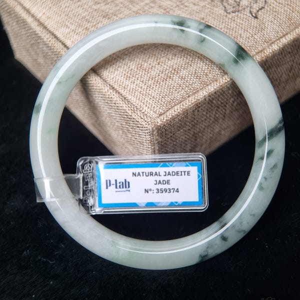 May include: A smooth, circular jadeite bangle featuring a translucent white base with deep green mottled inclusions. A small rectangular tag is attached to the bangle, displaying the text P-lab, NATURAL JADEITE JADE, and N: 359374.