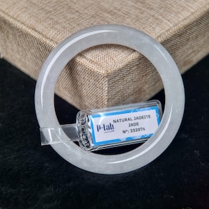 May include: A pale green jadeite bangle bracelet. The round bracelet has a smooth, polished surface. A tag attached to the bracelet reads "NATURAL JADEITE JADE" and "Nº: 332076". The bracelet is displayed on a dark surface.