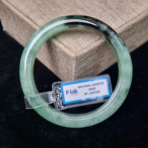 May include: A green jadeite bangle bracelet with a polished finish. The bracelet has a smooth surface with varying shades of green. A tag attached to the bracelet reads "NATURAL JADEITE JADE". The bracelet is displayed on a textured, brown surface.