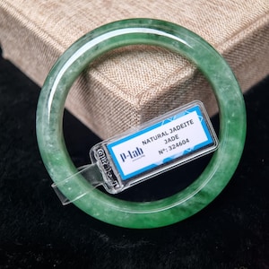 May include: A translucent green jadeite bangle bracelet. The bracelet is round and smooth, with variations in the green color. A tag with the text "NATURAL JADEITE JADE" and the number "324604" is attached to the bracelet.