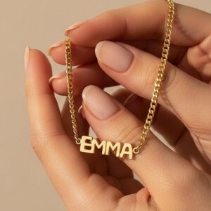 Op de afbeelding: Een goudkleurige ketting met de naam "EMMA" in blokletters. De ketting wordt vastgehouden door twee handen met verzorgde nagels. De ketting heeft een curb-stijl. De achtergrond is effen en neutraal.