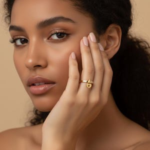 May include: A gold ring with a heart-shaped padlock charm. The ring has a band with small, clear stones and a dangling padlock pendant. The ring is worn on a finger.